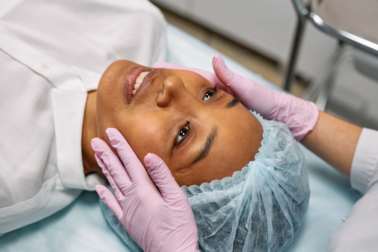 A woman receiving a facial treatment in a spa, wearing a hairnet while hands in gloves gently touch her face.